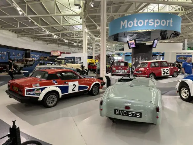 Een showroom in het British Motor Museum toont verschillende klassieke auto's, waaronder een rode raceauto met nummer 2, een groene cabriolet met kenteken MVC 575 en een rode auto met nummer 52. Boven hangt een bord "Motorsport" dat alle bezoekers tijdens hun bezoek kunnen bewonderen.