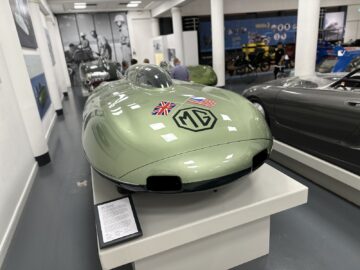 Een strakke groene MG-sportwagen tentoongesteld in het British Motor Museum, met Britse en Amerikaanse vlaggen op de motorkap en een informatief bord eronder. Perfect voor een bezoek of zelfs een reportage over klassieke voertuigen.