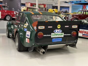 Achteraanzicht van een groene raceauto met meerdere sponsorstickers, tentoongesteld in een goed verlicht automuseum tijdens ons bezoek aan het British Motor Museum. Op de achtergrond zijn andere klassieke auto's zichtbaar.