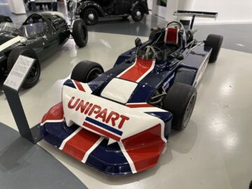 Een vintage raceauto met het "Unipart"-logo, versierd met een Union Jack-ontwerp op de neus, tentoongesteld in de showroom van het British Motor Museum. De auto heeft grote voor- en achtervleugels en slicks, waardoor hij een blikvanger is in iedere bezoek- of motorsportreportage.