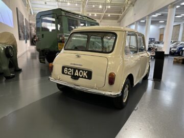 Binnen staat een klassieke crèmekleurige auto met kenteken "621 AOK", omringd door andere voertuigen in een museumachtige setting. Deze tentoonstelling kunt u ontdekken tijdens uw bezoek aan het British Motor Museum.