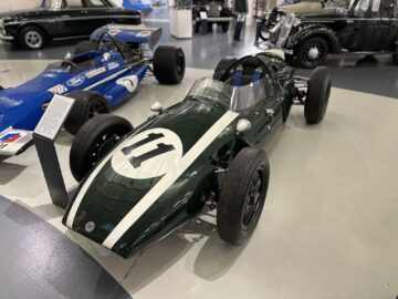 Een vintage racewagen met het nummer 11 op de motorkap wordt tentoongesteld in een overdekte showroom in het British Motor Museum. Het is donkergroen met witte racestrepen en omringd door andere klassieke en vintage auto's, wat zorgt voor een boeiend bezoek.