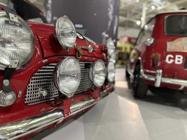 Close-up van twee klassieke rode auto's in het British Motor Museum, met de nadruk op de grilles aan de voorkant en meerdere koplampen op de auto op de voorgrond, met een deel van de achterkant van de tweede auto in zicht. Deze boeiende reportage geeft hun tijdloze charme perfect weer.