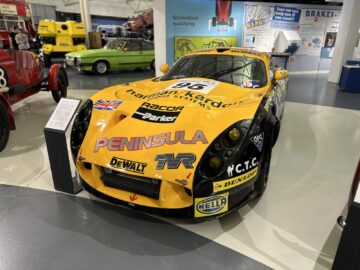 Geel-zwarte raceauto met verschillende sponsorlogo's tentoongesteld op een overdekte tentoonstelling in het British Motor Museum. Andere oldtimers en informatiedisplays zijn op de achtergrond zichtbaar en bieden een rijke ervaring voor bezoekers die geïnteresseerd zijn in de autogeschiedenis.