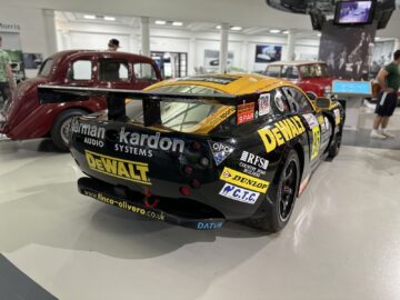 Een zwart-gele raceauto met DEWALT-logo te zien in het British Motor Museum. Bezoekers kunnen ook genieten van een reportage van andere oldtimers op de achtergrond, waardoor het een fascinerend bezoek is voor autoliefhebbers.