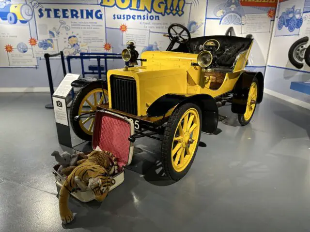 Een geel-zwarte vintage auto is te zien in het British Motor Museum. Ernaast staat een klein, open koffertje met knuffels, waaronder een tijger en een pop. Op de achtergrond zijn informatiepanelen zichtbaar, waardoor dit museumbezoek het gevoel krijgt dat u in een nostalgische reportage stapt.