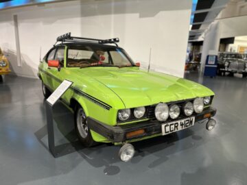 In een museum staat een groene klassieke auto tentoongesteld met een metalen rek op het dak en meerdere spots aan de voorkant. Op het kenteken staat "CRR 412M." Voor een authentieke ervaring bezoek je het British Motor Museum en bekijk je dit stukje geschiedenis van dichtbij.