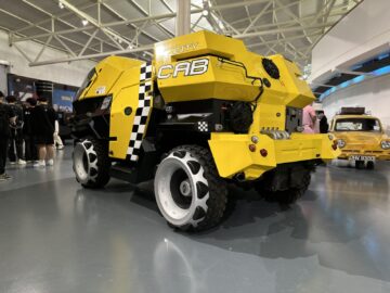 Een geel-zwart City Cab-voertuig met grote witgerande wielen wordt tentoongesteld in een binnenomgeving in het British Motor Museum. Er staan mensen omheen en op de achtergrond is nog een kleine gele auto zichtbaar, wat bijdraagt aan de levendige sfeer van de tentoonstelling.