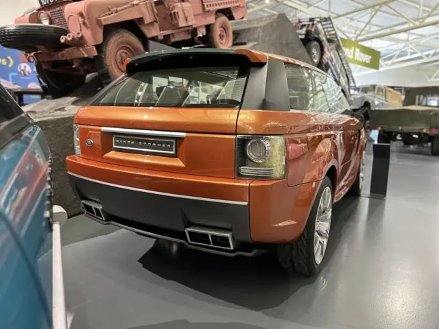 Een feloranje Range Rover SUV wordt binnen tentoongesteld in het British Motor Museum. Andere voertuigen, waaronder een ouder model, zijn op de achtergrond zichtbaar. Bezoek deze indrukwekkende tentoonstelling voor een fascinerende autoreis.