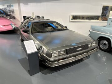 Een DeLorean DMC-12 tentoongesteld in het British Motor Museum, met roestvrijstalen carrosseriepanelen en vleugeldeuren. Naast het voertuig staat een informatiebord dat bezoekers een gedetailleerde reportage over de geschiedenis ervan biedt.