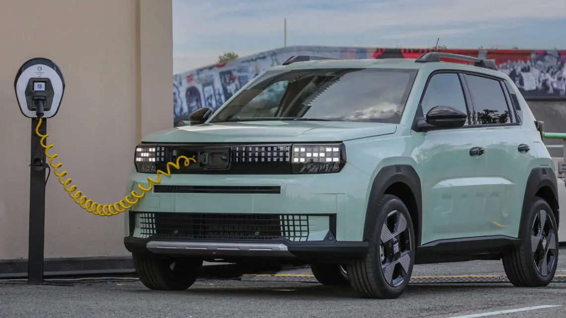 Un SUV eléctrico de color verde pálido está enchufado a una estación de carga en el exterior, mostrando su perfil frontal y lateral, que recuerda al elegante Fiat Grande Panda con sus exclusivas cadenas cinemáticas.