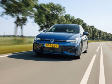 Een blauwe VW Golf met het Nederlandse kenteken "GDT-83-L" rijdt op een zonnige dag over een met bomen omzoomde weg.