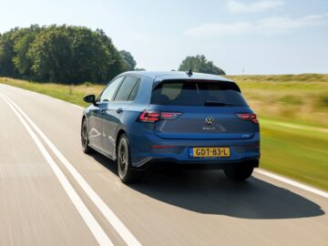 A blue VW Golf drives on a two-lane road through a picturesque landscape with trees and green fields on a sunny day. The license plate reads 