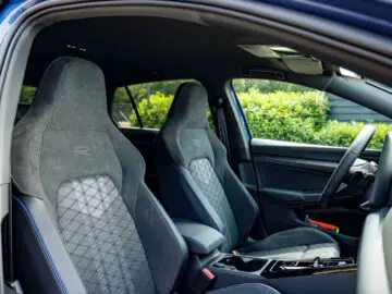 Interior of a modern VW Golf with two front seats with diamond-patterned upholstery, a steering wheel and part of the dashboard. The car door is open and green bushes appear outside.