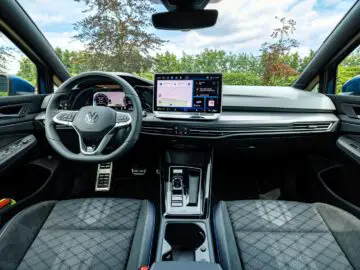 Binnenaanzicht van een VW Golf-voertuig met het stuur, het dashboard met digitale displays en de middenconsole met versnellingspook. De achtergrond bestaat uit bomen en een gedeeltelijk bewolkte hemel.