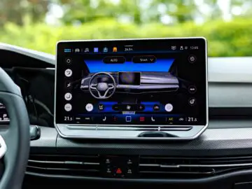 Close-up of the infotainment screen of a VW Golf showing the climate control interface and various options, with a green background visible through the windows.