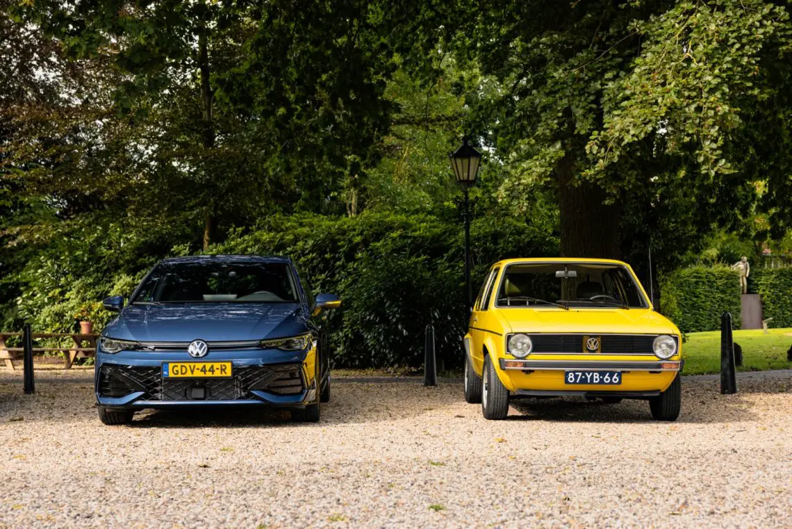 Een moderne blauwe VW Golf en een vintage gele Volkswagen-auto staan naast elkaar geparkeerd op een grindoppervlak met bomen op de achtergrond.