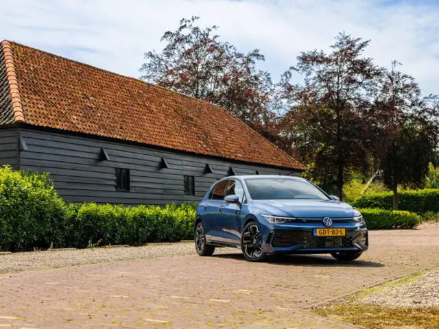 Een blauwe VW Golf-hatchback staat geparkeerd op een geplaveide oprit voor een rustiek houten gebouw met een rood pannendak. Struiken en bomen omringen het gebied onder een licht bewolkte hemel.