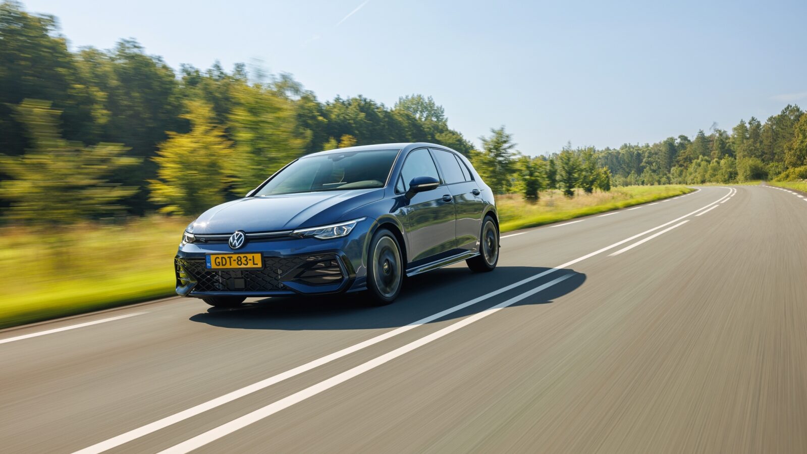 Een VW Golf in levendig blauw rijdt langs een zonnige, met bomen omzoomde landelijke weg.