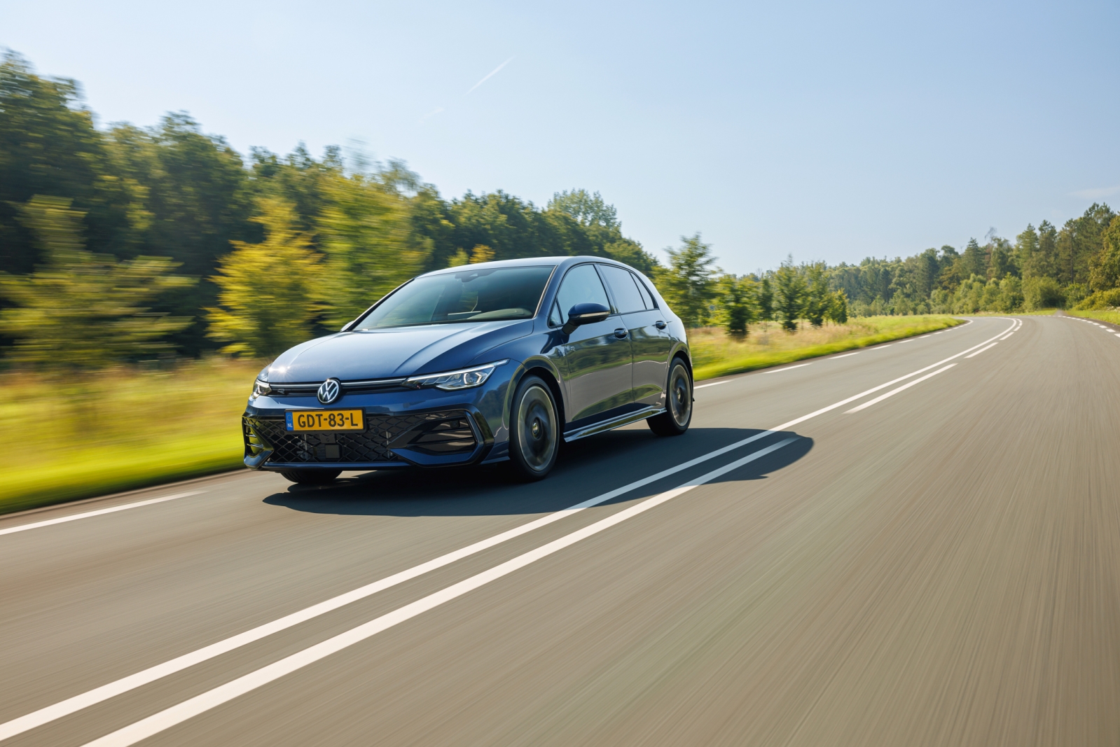 Een VW Golf in levendig blauw rijdt langs een zonnige, met bomen omzoomde landelijke weg.