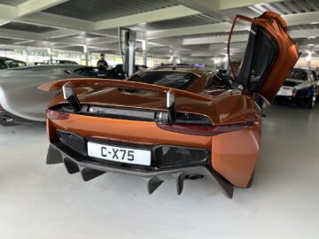 A rear view of an orange sports car with the left door open in a showroom. The Jaguar Heritage Trust Collection piece has a license plate reading 