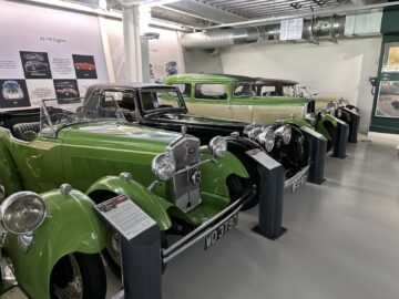 A row of vintage green cars from the Jaguar Heritage Trust collection is displayed in a museum, each with informative signs in front of it. Posters and diagrams of car engines can be seen in the background, providing a detailed account of automotive history.