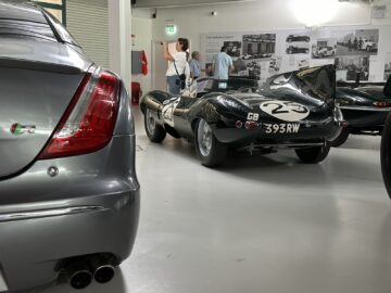 A classic race car number 2 and license plate 393 RW, part of the Jaguar Heritage Trust Collection, is displayed in an exhibition. In the background, two people are talking. The back of a gray modern car is visible in the foreground.