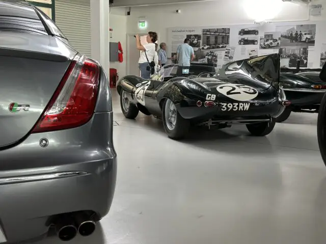 Een klassieke raceauto met nummer 2 en kenteken 393 RW, onderdeel van de Jaguar Heritage Trust Collection, wordt tentoongesteld in een tentoonstelling. Op de achtergrond zijn twee mensen aan het praten. De achterkant van een grijze moderne auto is zichtbaar op de voorgrond.