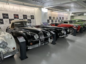 A row of classic cars, featuring black and white vintage models, is displayed in an indoor car exhibition with information panels on the wall behind them documenting their rich history. Among these treasures is a piece from the Jaguar Heritage Trust Collection.