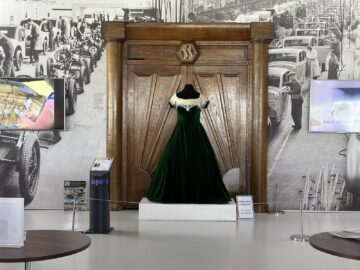 A green velvet dress displayed in front of a large wooden gate, with a vintage photo of an auto assembly line from the Jaguar Heritage Trust Collection in the background. Set up around the exhibit are tablets and information panels that offer detailed reports.