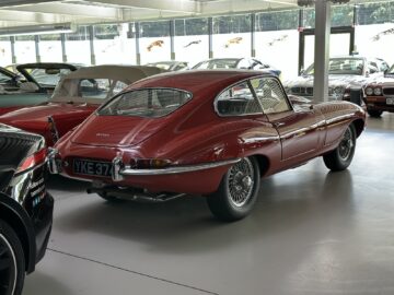 A red vintage Jaguar E-Type car, part of the prestigious Jaguar Heritage Trust collection, is parked inside a showroom surrounded by an eclectic mix of classic and modern cars.