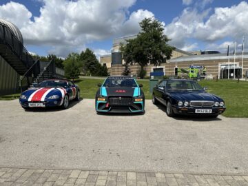 On a sunny day, three parked cars are displayed outside: a sports coupe with racing stripes on the left, a branded race car from the Jaguar Heritage Trust Collection in the middle and a classic sedan on the right, with buildings in the background.