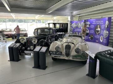 An exhibition of vintage cars, including a black model with license plate CDU 700 and a white model with license plate NUB 120, from Jaguar Heritage Trust's collection, in a museum setting with an exhibit on influential women in automotive history.