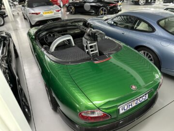 A green convertible sports car with a large machine gun mounted on the passenger seat, parked in a showroom alongside other luxury vehicles, features prominently in the Jaguar Heritage Trust Collection.