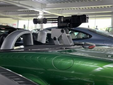 A green car with mounted machine gun is displayed in an indoor showroom, surrounded by other vehicles from the Jaguar Heritage Trust Collection.