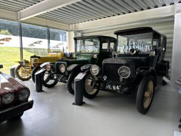 A report-worthy display of vintage cars, including a green car, a black car and a yellow car from the Jaguar Heritage Trust Collection, is carefully arranged in a well-lit showroom with large windows.