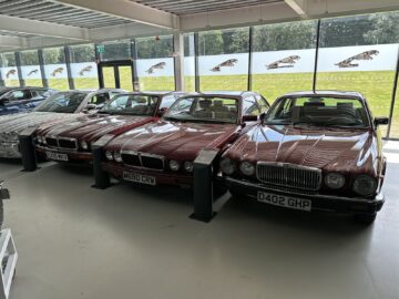 A row of classic Jaguar cars from the Jaguar Heritage Trust Collection is displayed in a showroom, with large windows in the background showing a lawn and iconic Jaguar logos.