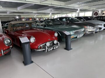 A range of cars on display in an indoor showroom includes a red classic convertible with a beige hood, a green Jaguar convertible from the Jaguar Heritage Trust collection, a silver sedan and a black car.