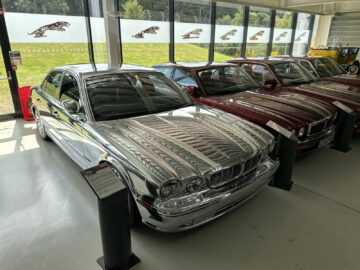 A collection of cars displayed indoors, with a chrome Jaguar Heritage Trust sedan in the foreground.