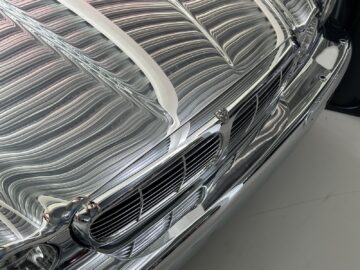 Close-up of the highly reflective, metal grille and hood on the front of a car under bright overhead lighting. The reflections create a complex pattern on the surface, reminiscent of the classic models preserved in the Jaguar Heritage Trust Collection.