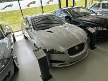 A silver Jaguar sports car displayed in an indoor showroom, part of the exclusive Jaguar Heritage Trust collection, flanked by other vehicles. Information boards were placed in front and beside the car.