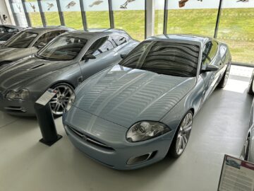 A light blue sports car, part of the Jaguar Heritage Trust Collection, is displayed indoors next to a silver car. A glass wall with emblems of different car models can be seen in the background, adding to the REPORTAGE aesthetic of the exhibition.