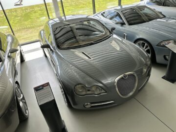 Inside, a sleek silver car is on display, part of the Jaguar Heritage Trust Collection. The polished vehicle stands by a large window surrounded by other cars. Information boards offer detailed reports on the history and significance of each model.