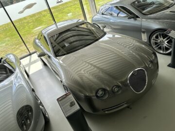 A sleek, silver luxury car from the Jaguar Heritage Trust Collection is displayed inside alongside other similar models. Large background windows let in natural light, highlighting the car's smooth, polished surface, creating a stunning scene worthy of premium reportage.