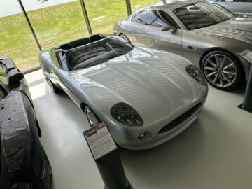 A sleek, silver convertible from the Jaguar Heritage Trust collection is displayed in a covered showroom, with informative signs next to it and other cars parked nearby. Large windows reveal a green landscape outside, perfectly framing this feat of automotive excellence.