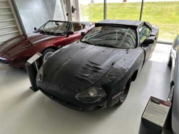 Two cars are displayed in a showroom. The car in the foreground, part of the Jaguar Heritage Trust Collection, is covered in a black material with visible wrinkles and holes. The other car, on the left, is a red convertible. Both cars are behind barriers secured with rope. This scene feels like car shooting at its best.