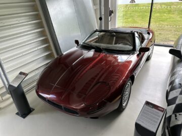 Inside, a maroon convertible is on display, with a sign and information boards next to it. Part of the Jaguar Heritage Trust collection, the vehicle has a sleek design and polished surface, reflecting the essence of classic automotive elegance.