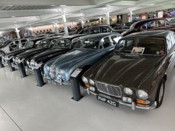 A row of vintage cars displayed indoors, including a brown car in the front with the imprint 'PHP 42G' and a light blue car next to it. Detailed information boards for each vehicle in this Jaguar Heritage Trust Collection provide rich historical reportage.