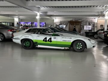 A vintage race car with number 44 on the side is parked in an indoor car show area, with other cars in the background. With its white and green livery, this classic beauty is a proud part of the Jaguar Heritage Trust Collection.