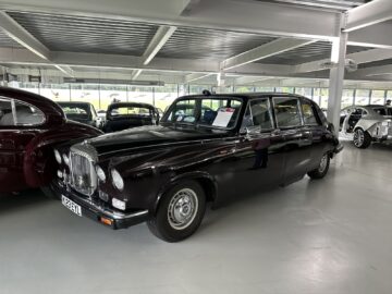 A classic black car is displayed in an indoor showroom, part of the Jaguar Heritage Trust collection, with a sign on the windshield detailing the vehicle. Other cars are visible in the background.